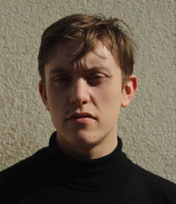 Outdoor headshot of Conor Booker wearing a black turtle neck shot against a textured cream wall