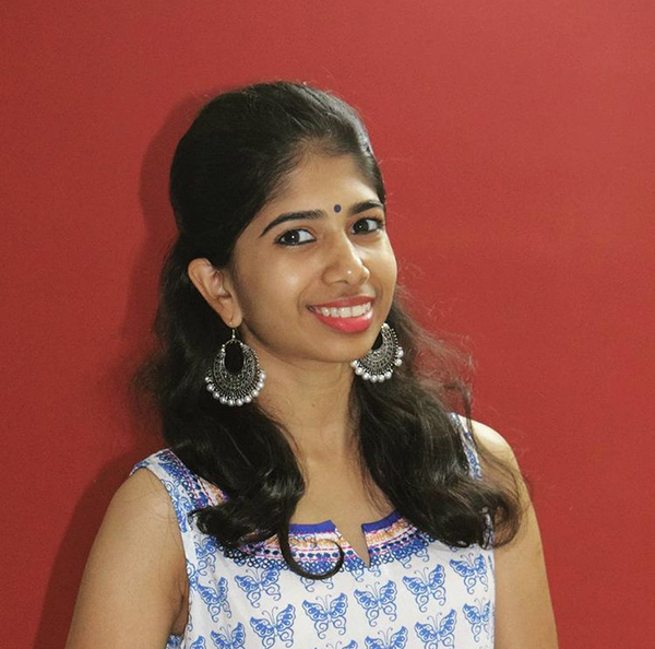 Indoor headshot of Divija Melally wearing a white and blue patterned top taken against a red background