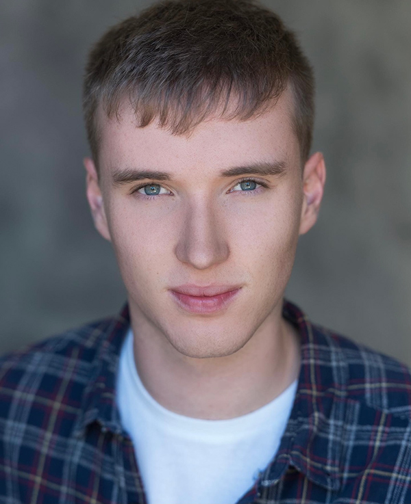 Studio headshot of Ryan Carroll wearing a white t shirt with a blue checked shirt layered over the top