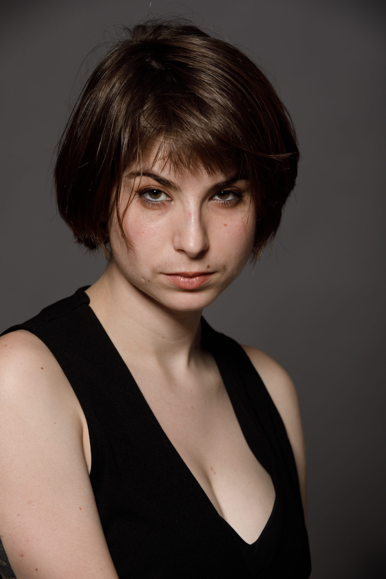 Headshot of artist with neck-length brown hair, dark eyes and a v-neck black top. Dark backdrop behind.