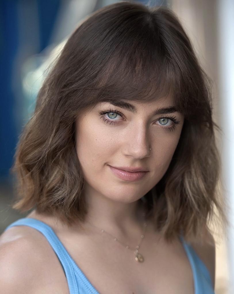 Headshot of female actress with should-length brown hair, blue eyes, wearing a necklace